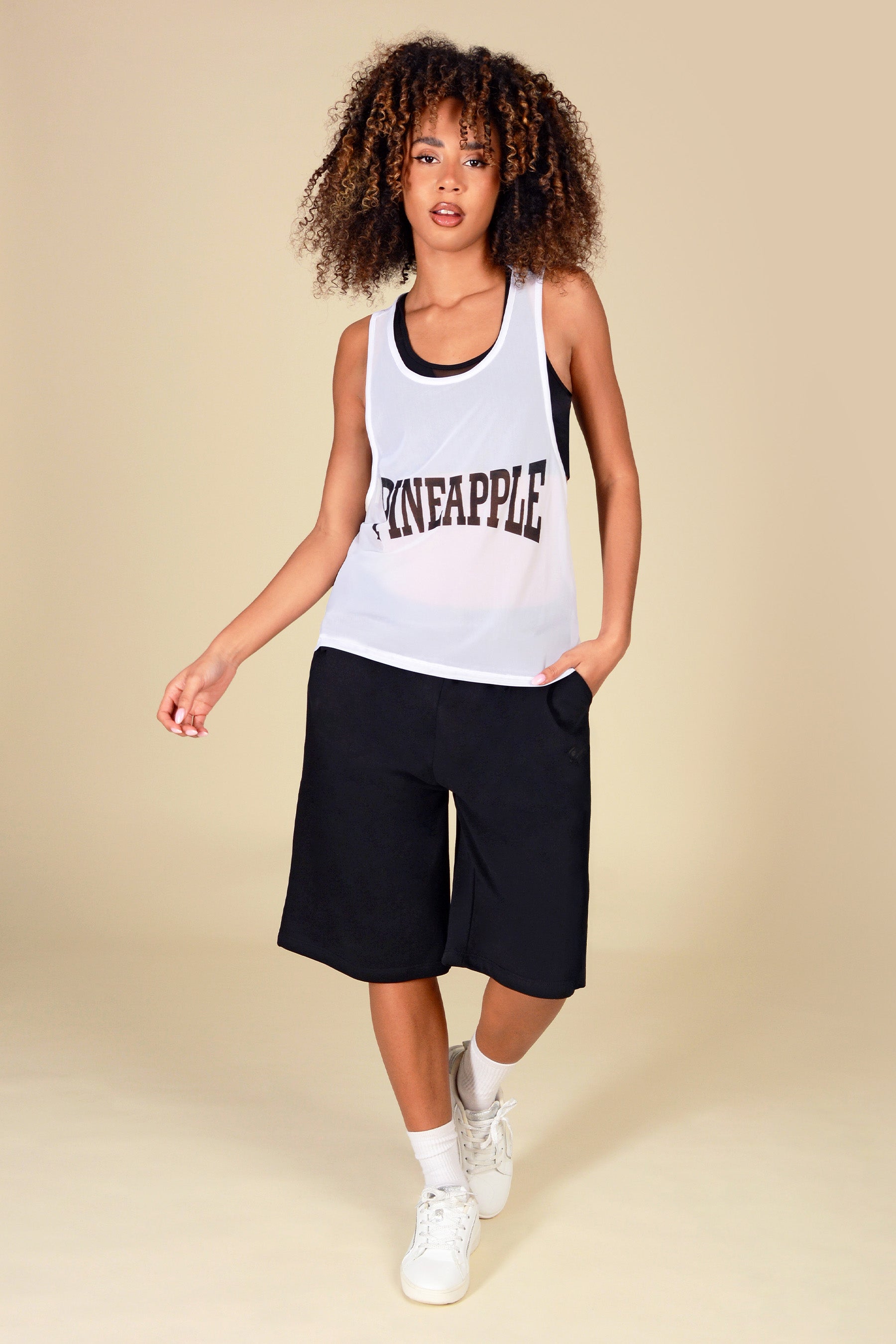 Woman wearing a white longline mesh  vest with 'Pineapple' print and black shorts on a beige background